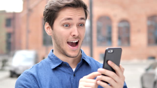 Outdoor-Portrait-of-Young-Man-Excited-for-Success-on-Smartphone
