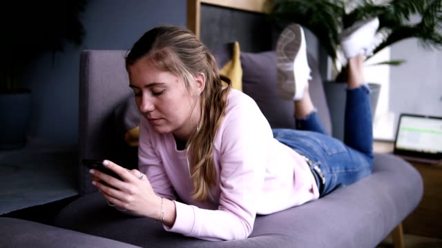 Attractive-girl-in-pink-sweater-using-smart-phone-while-lying-on-the-couch-in-the-living-room.-Typing,-watching-video,-or-browsing-social-media-on-smartphone.-In-front-her-laptop.-Slow-motion.-Front-view