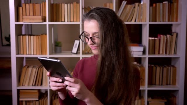 Retrato-de-primer-plano-de-joven-estudiante-caucásica-en-gafas-usando-la-tableta-sonriendo-y-mirando-la-cámara-en-el-interior-de-la-biblioteca-de-la-universidad-en-el-interior