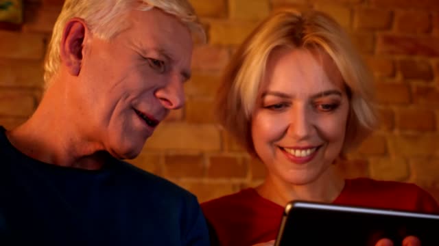 Closeup-shoot-of-aged-happy-couple-using-the-tablet-sitting-on-the-sofa-resting-together-indoors-in-a-cozy-apartment