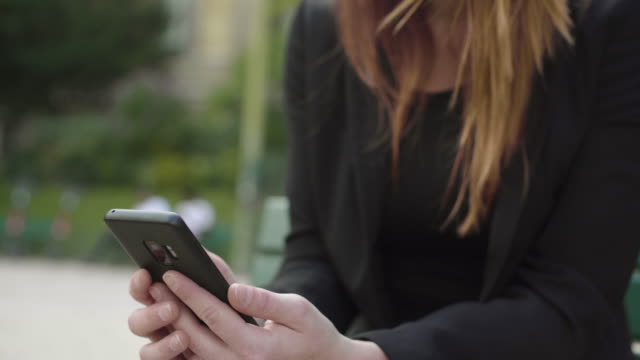 Geschäftsfrau-mit-Smartphone-Sitzen-auf-der-Bank-im-Park-in-Paris,-lachen,-während-SMS-mit-Handy-draußen,-Tippen-von-SMS-auf-Handy.-Inhalt,-rote-Haare.-soziale-medien.-lächelnd.-4K.