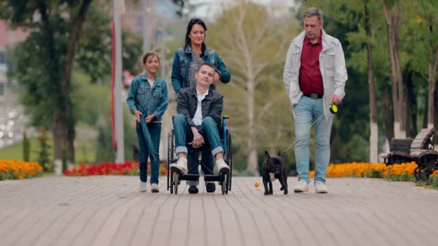 Guy-im-Rollstuhl-läuft-mit-seiner-Familie-die-Allee-entlang.-Familienförderung-für-Menschen-mit-Behinderungen.