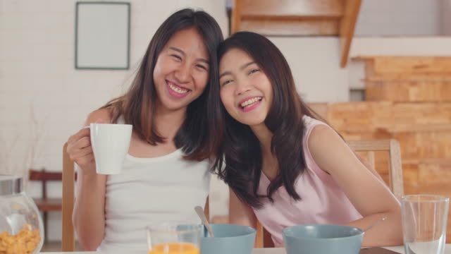 Asiatische-Lesbische-paar-glücklich-zu-schüchternlächeln,-die-in-die-Kamera-schauen,-während-Sie-morgens-in-der-Küche-frühstücken.