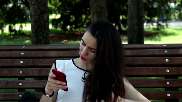 Retrato-de-cerca-Una-hermosa-joven-con-una-camiseta-blanca-está-chateando-en-las-redes-sociales-en-su-teléfono-inteligente-mientras-está-sentada-en-un-banco-en-un-parque.