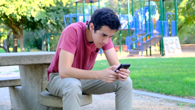 Joven-usando-su-teléfono-inteligente-sentado-en-un-banco-del-parque