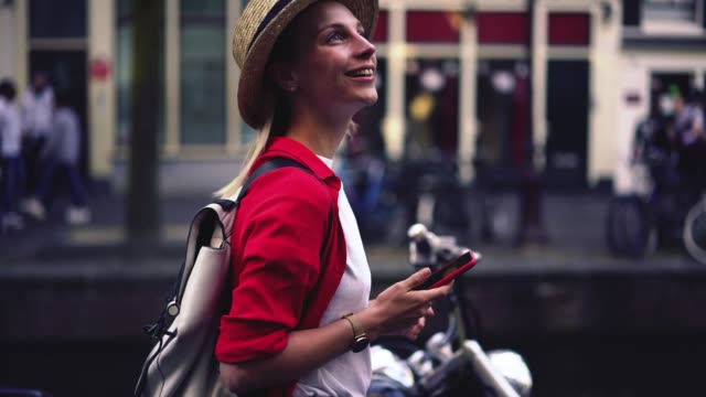 Side-view-of-smiling-Caucasian-hipster-girl-in-casual-wear-and-hat-walking-outdoors-on-city-street-and-holding-mobile-phone-in-hands