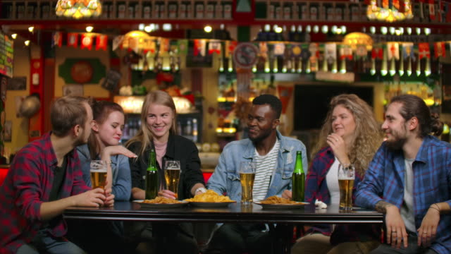 African-American-with-friends-at-the-bar-drinks-beer-and-eats-chips,-friends-raise-glasses-and-bottles-and-knock-/-check-on-the-table-smiling-and-saying-toasts