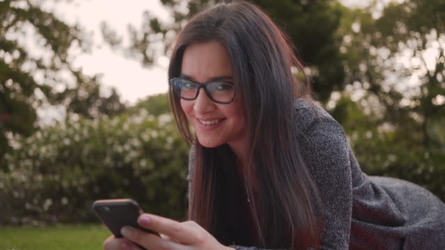 Portrait-of-a-beautiful-young-woman-wearing-eyeglasses-lying-on-park-looking-to-camera-while-using-smartphone-in-the-park
