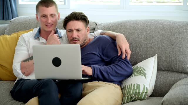 Gay-couple-relaxing-on-couch-using-laptop-computer.-Interesting-mood.
