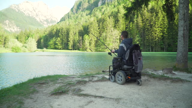 Resolución-4k-de-pescador-discapacitado-en-una-silla-de-ruedas-eléctrica-pescando-en-un-hermoso-lago-cerca-del-bosque-y-la-montaña-en-la-parte-posterior
