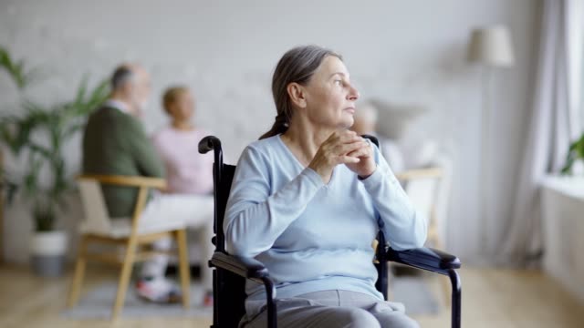 Tracking-shot-of-disabled-senior-woman-in-wheelchair-looking-away-and-contemplating-in-nursing-home,-other-aged-patients-in-background