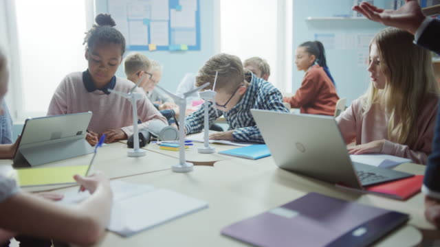 In-der-Grundschule:-Klasse-der-brillanten-kleinen-Kinder-arbeiten-als-Team-mit-Tablet-Computern,-um-Windkraftanlagen-zu-programmieren.-Klassenzimmer-mit-Kindern-Lernen-über-umweltfreundliche-Formen-erneuerbarer-Energie