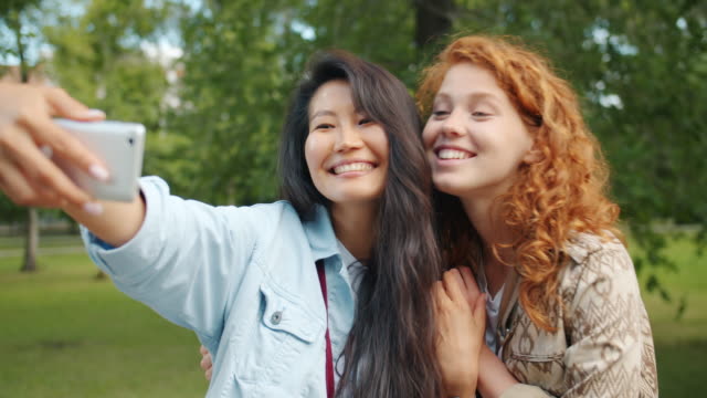 Chicas-lindas-tomando-selfie-con-el-teléfono-inteligente-posando-en-el-parque-verde-abrazando-sonriendo