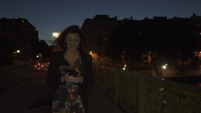 Happy-caucasian-modern-woman-wearing-flower-dress,-black-jacket-and-red-hair-walking-through-the-street-and-writing-a-text-message-on-her-smartphone-by-night.-Paris-4K-UHD.-Slow-Motion.-Medium-Shot.