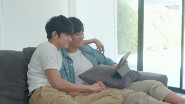 Young-Gay-couple-using-tablet-at-home.-Asian-LGBTQ-men-happy-relax-fun-using-technology-watching-movie-in-internet-together-while-lying-sofa-in-living-room-concept.-Slow-motion-Shot.