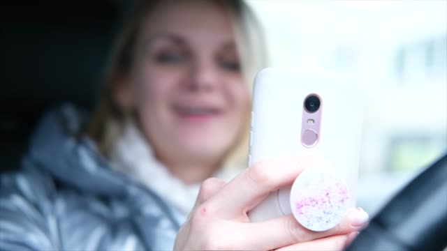 Blurred-blonde-woman-blogger-in-winter-clothes-sitting-in-car-and-talking-with-followers,-live-streaming,-looking-to-smartphone-screen.-focus-on-phone