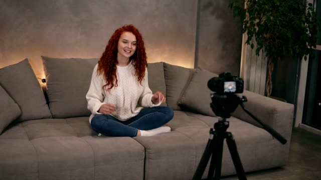 Attractive-curly-red-headed-young-girl-vlogger-is-talking-in-front-of-camera-recording-video-for-online-blog-in-internet-speaking,-smiling.-Woman-is-wearing-jeans-and-white-sweater