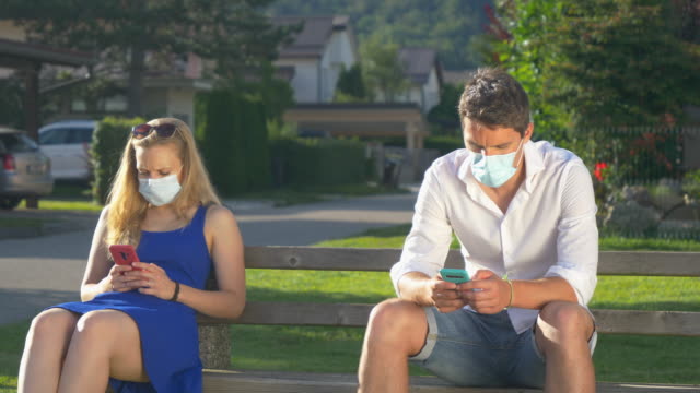 CLOSE-UP:-Two-strangers-wear-facemasks-while-sitting-on-park-bench-and-texting.