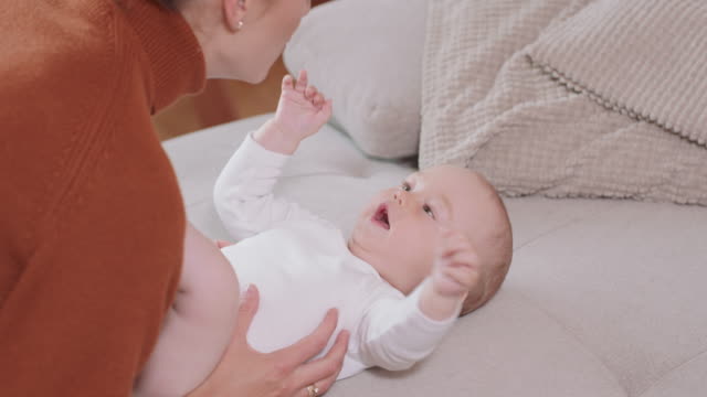 Close-up-of-the-baby-lying-on-its-back-on-the-couch,-smiling-and-happy,-shaking-hands,-looking-at-its-mother,-who-kneels-by-the-baby,-talks-to-it,-tickels-and-smiles