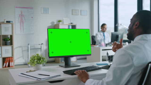 African-American-Medical-Doctor-macht-einen-Videoanruf-mit-Patient-auf-einem-Computer-mit-Green-Screen-Display-in-einer-Gesundheitsklinik.-Assistant-in-Lab-Coat-spricht-über-Gesundheitsprobleme-im-Krankenhausbüro.