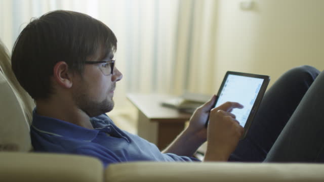 Man-is-Laying-on-Couch-and-Typing-a-Message-on-Tablet-at-Home