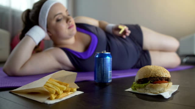 Lazy-lady-in-sportswear-lying-on-yoga-mat-eating-junk-food-instead-of-training