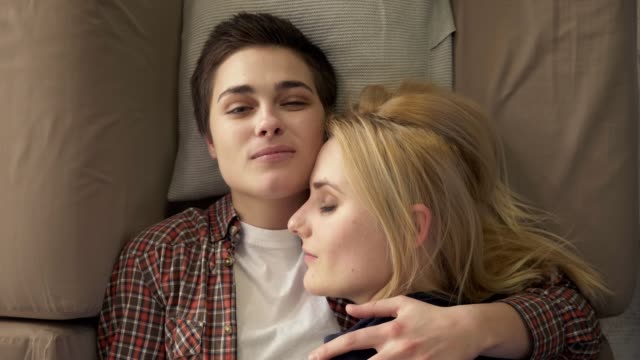 Two-young-lesbian-girls-lie-on-the-couch,-hug,-cuddle,-sleep,-girl-with-short-hair-looks-at-the-camera,-lgbt-family-concept,-happy,-top-shot,-relaxation-60-fps