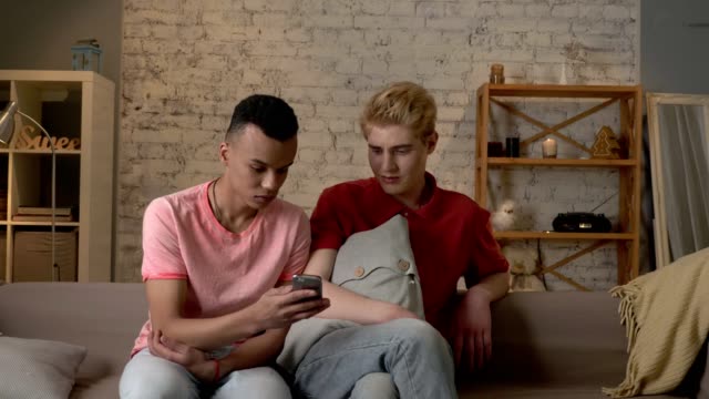 Two-international-friends-of-a-homosexual-are-sitting-on-the-couch-and-watching-disgusting,-mercenary-pictures-on-a-smartphone.-Home-cosiness,-family,-internet-concept.-60-fps