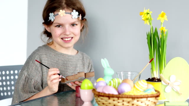 A-brown-eyed-girl-with-a-wreath-on-her-head-is-preparing-for-Easter.-The-child-looks-at-the-camera-and-laughs-cheerfully.-In-her-hands-paint-and-brush.-On-the-table-is-an-Easter-basket-and-a-bouquet-of-daffodils