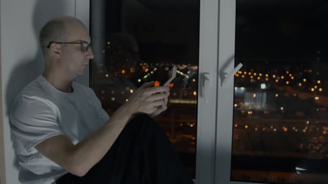Glatzkopf-in-Gläser-Suche-Tablet-pc-sitzt-auf-der-Fensterbank-mit-Nacht-Stadtbild
