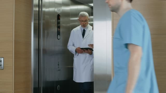 In-the-Hospital-Two-Doctors-Walk-out-of-the-Elevator-while-have-Discussion-while-Using-Tablet-Computer.-In-the-Background-Patients-and-Medical-Personnel.-New-Modern-Fully-Functional-Medical-Facility.