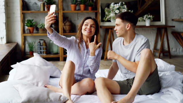 Modern-married-couple-is-taking-selfie-in-bedroom-gesturing-posing-and-kissing-while-sitting-on-bed-together.-Modern-technology-and-people-concept.