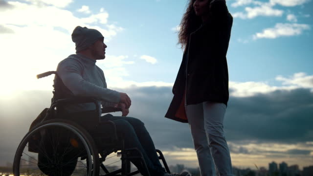 Young-woman-with-disabled-man-in-a-wheelchair-talking-outdoors-at-sunset