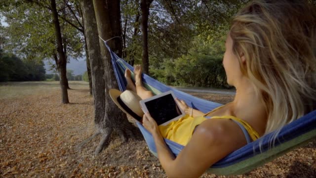 Frau-in-Hängematte-lesen-auf-digital-Tablette-mit-mobilen-Anwendungen-auf-wireless-Technologie.-Junge-Frau,-die-Natur-genießen,-im-Spätsommer-pendeln-zwischen-Bäumen
