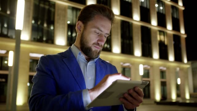 Panning-medium-shot-of-handsome-bearded-businessman-in-blue-jacket-using-tablet-computer-in-night-illuminated-city-street