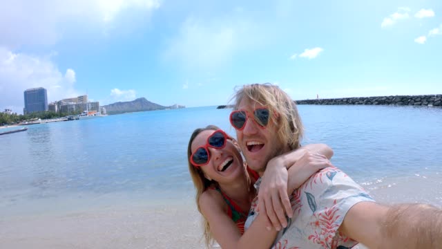 Couple-taking-selfies-on-Waikiki-Beach-in-Honolulu,-Hawaii.-Selfie-point-of-view-wide-angle-of-Waikiki-Beach.-Young-couple-taking-selfies-with-heart-shaped-sunglasses