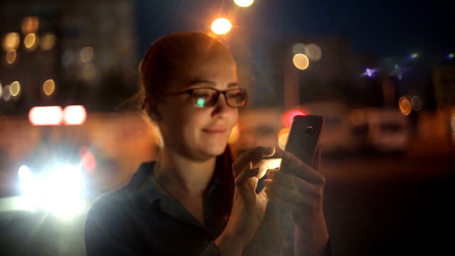 Retrato-de-la-joven-mujer-caucásica-hermosa-usando-la-mano-del-teléfono-inteligente-mantener-al-aire-libre-en-la-noche-de-la-ciudad,-sonriendo,-cara-iluminada-ScreenLight-red-social,-tecnología,-concepto-de-comunicación