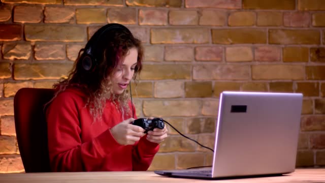 Portrait-of-young-female-blogger-in-red-hoodie-playing-video-game-using-laptop-and-joystick-on-bricken-wall-background.