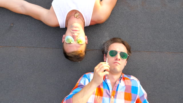 Top-view-of-young-man-lying-with-his-boyfriend-on-roof-and-giving-a-cigarette-to-him.-Handsome-male-couple-spending-time-on-rooftop-of-high-rise-building-and-smoking-together.-Close-up-Slow-motion