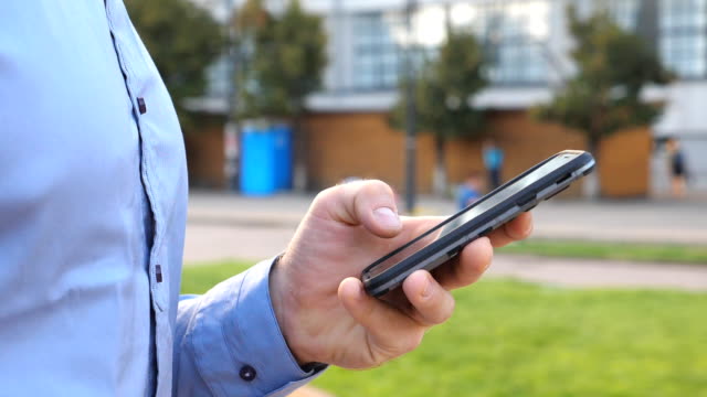 Close-up-hand-of-young-businessman-holding-and-touching-smartphone.-Unrecognizable-man-standing-on-urban-street-and-browsing-news-at-the-phone.-Guy-using-gadget-for-work.-Slow-motion-Dolly-shot