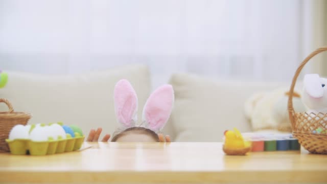 Cute-little-girl-with-bunny-ears-is-hiding-under-the-table-full-of-Easter-decorations.-Little-cute-white-girl-is-looking-with-amusement.-Laught-is-in-the-room.