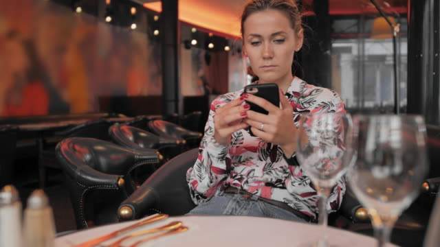 Atractiva-mujer-caucásica-utiliza-un-teléfono-móvil-mientras-se-sienta-en-un-restaurante.-El-concepto-de-esperar-un-pedido.-Slow-Motion