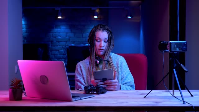 Closeup-shoot-of-young-attractive-female-blogger-with-dreadlocks-in-headphones-streaming-live-using-the-tablet-with-the-neon-background-indoors