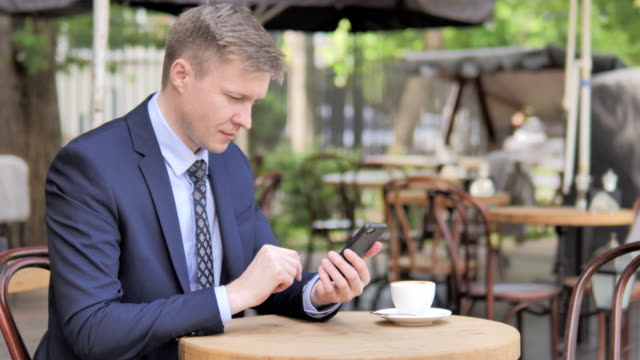 Businessman-Celebrating-Success-on-Phone,-Sitting-in-Outdoor-Cafe