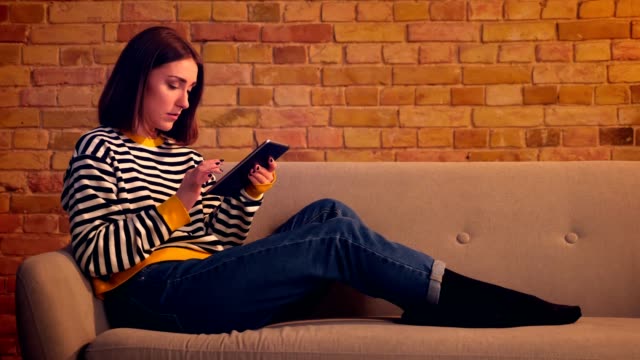 Closeup-profile-portrait-of-young-pretty-girl-using-the-tablet-sitting-on-the-sofa-in-a-cozy-apartment-indoors