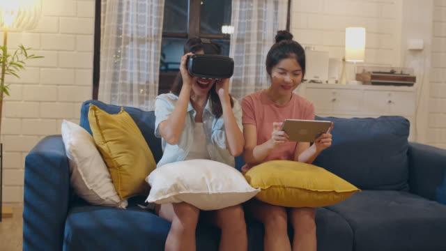 Lesbian-lgbt-women-couple-using-tablet-at-home,-Asian-female-feeling-happy-using-laptop-and-VR-playing-games-together-while-lying-sofa-in-living-room-in-night.-Lover-celebrate-holiday-concept.