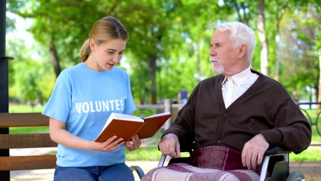 Young-female-volunteer-reading-book-for-disabled-pensioner-in-wheelchair,-care