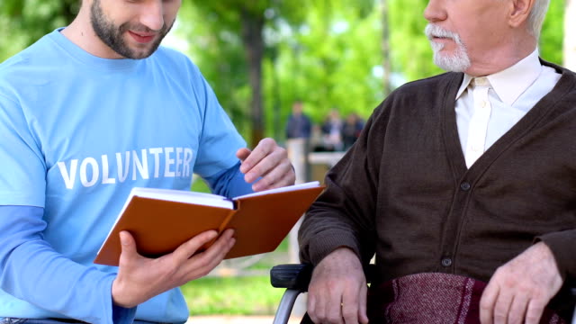 Freundliche-männliche-Freiwillige-Lesen-Buch-zu-lächeln-alten-Mann-im-Rollstuhl,-Hilfe