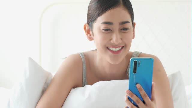 Mujer-asiática-hablando-en-tiempo-real-con-el-teléfono-inteligente-en-la-habitación-de-la-cama.-Concepto-de-tecnología,-comunicación-y-redes-sociales.