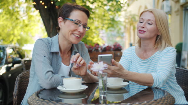 Cheerful-ladies-expressing-positive-emotions-using-smartphone-in-cafe-outdoors